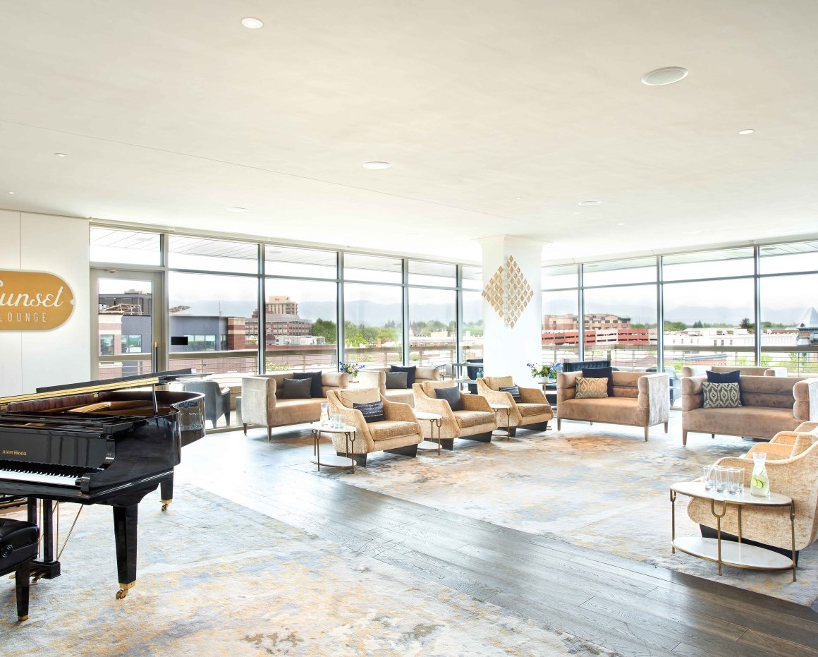 The dining room of the Sunset Lounge at The Elizabeth, with a black grand piano adjacent to a nook of comfortable chairs and chaises, by two bright bay windows