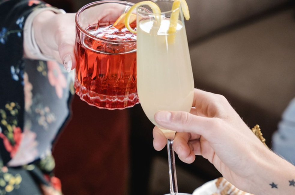 Two people toasting with vibrant cocktails, one red and one pale yellow, in a celebratory moment.