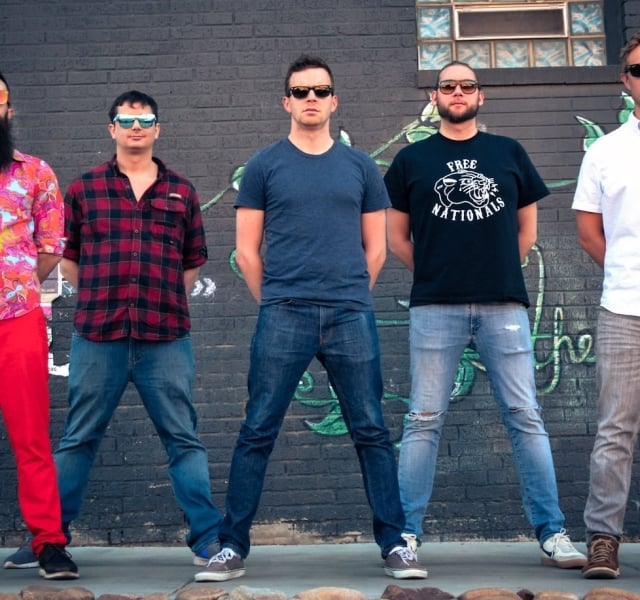 A five-man band stands on a stage with blue lighting behind them.