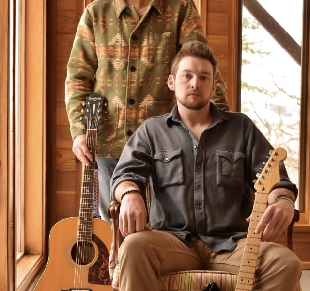 Musicians with a beard, wearing a hat, stands in a room with acoustic guitars next to another musician who is sitting