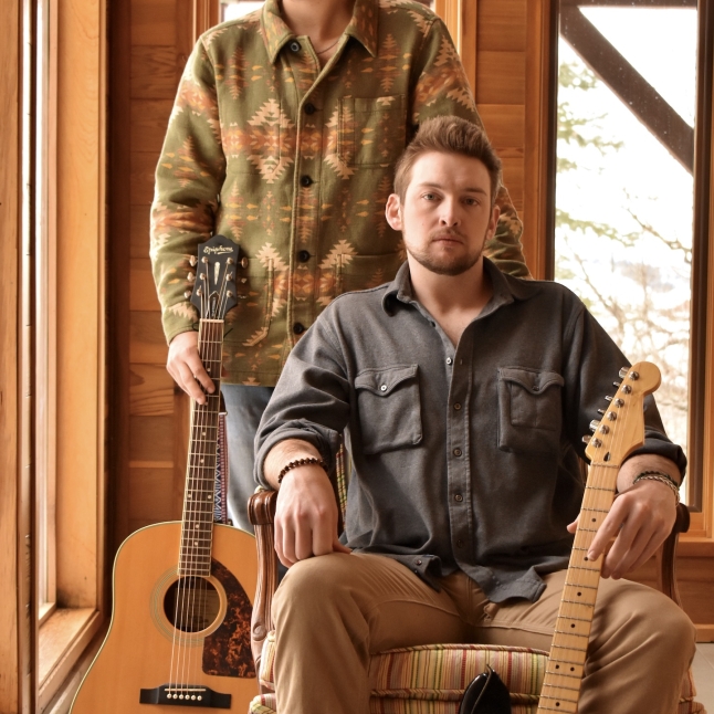 Musicians with a beard, wearing a hat, stands in a room with acoustic guitars next to another musician who is sitting