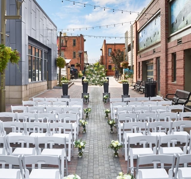 An outdoor wedding ceremony setup in a wide, urban alleyway flanked by brick buildings, with rows of white chairs facing the altar.