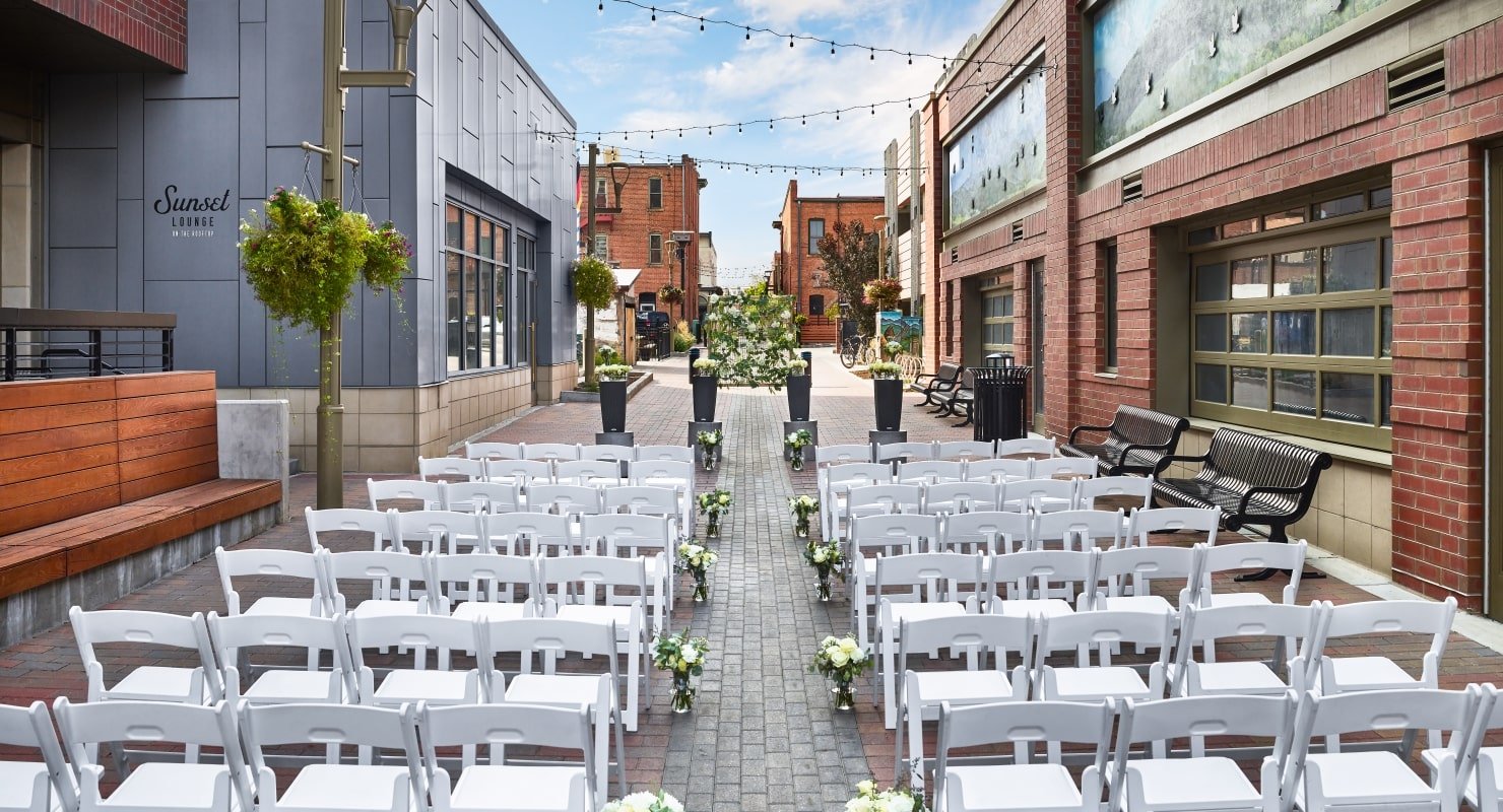 An outdoor wedding ceremony setup in a wide, urban alleyway flanked by brick buildings, with rows of white chairs facing the altar.