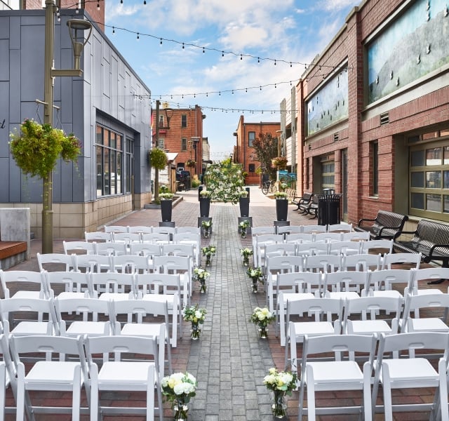 An outdoor wedding ceremony setup in a wide, urban alleyway flanked by brick buildings, with rows of white chairs facing the altar.