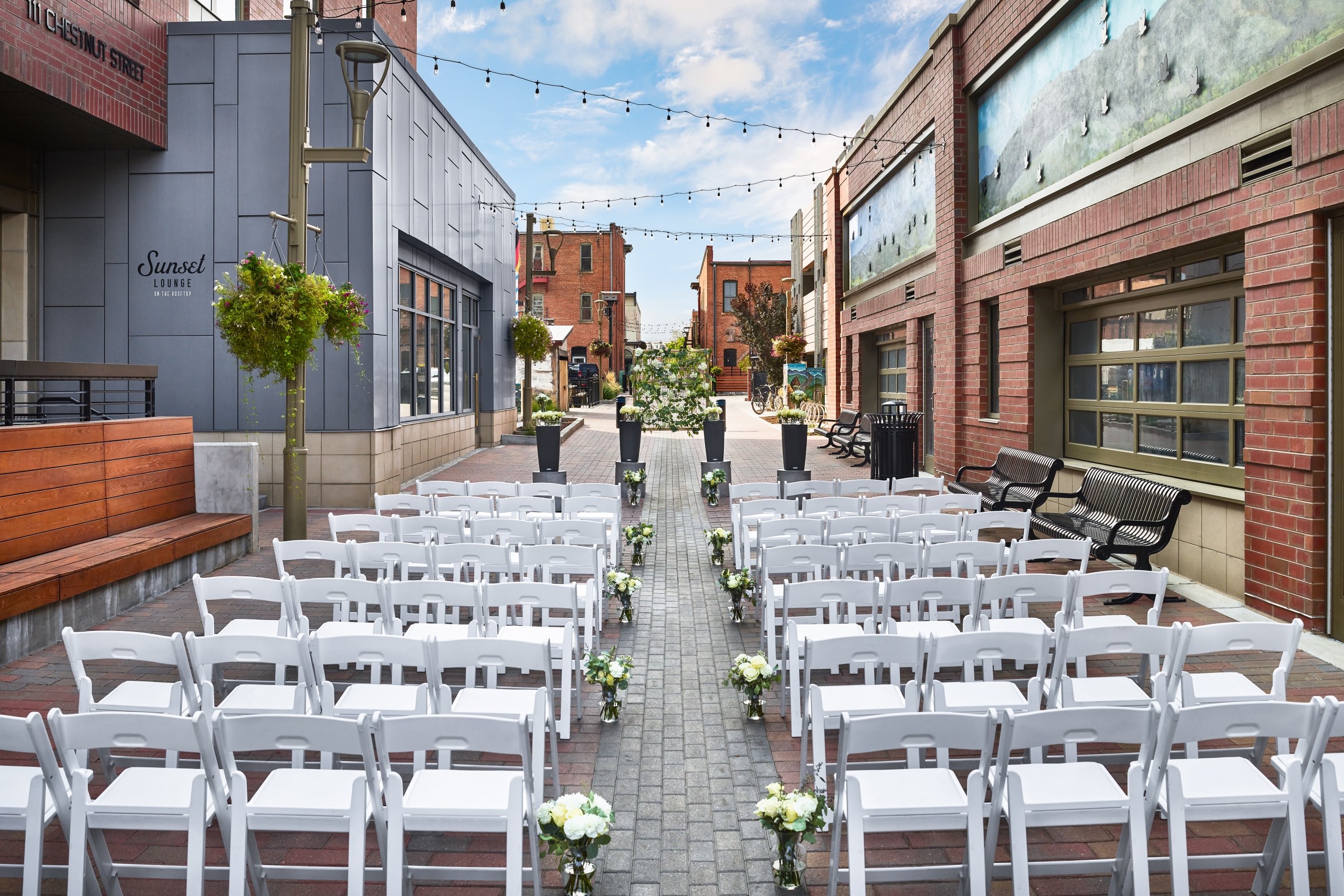 An outdoor wedding ceremony setup in a wide, urban alleyway flanked by brick buildings, with rows of white chairs facing the altar.