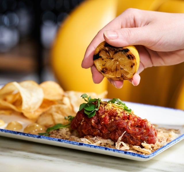 A hand squeezes burnt lemon onto a saucy chicken dish