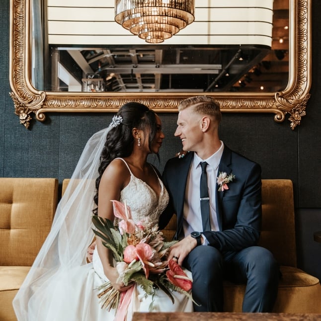 A bride holding a bouquet and groom seated on a gold fabric bench, under a large mirror, stare deeply into each others' eyes