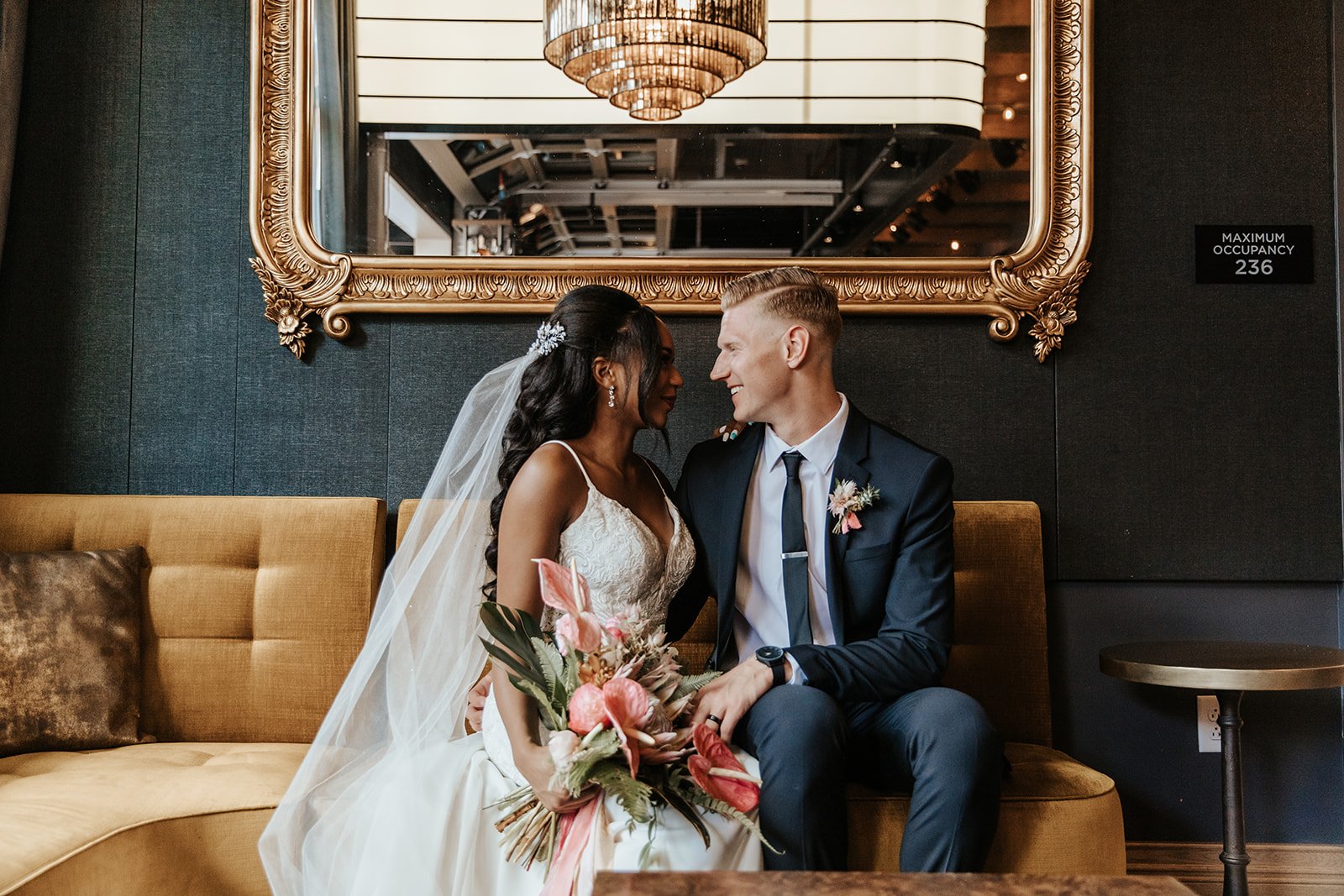 A bride holding a bouquet and groom seated on a gold fabric bench, under a large mirror, stare deeply into each others' eyes