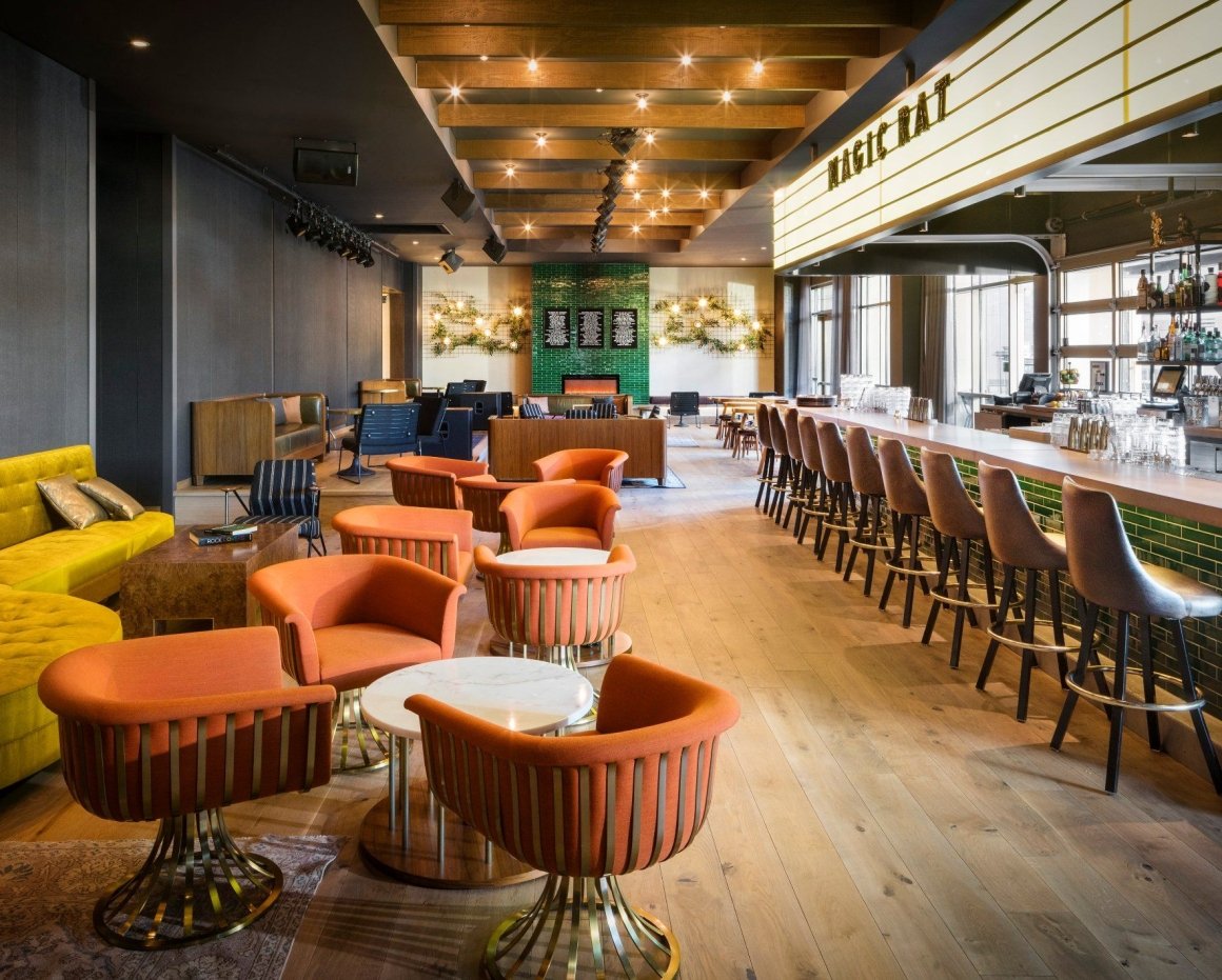 The bar at Magic Rat, a live music venue in the Elizabeth Hotel, featuring warm recessed lighting, a long bar and retro orange chairs around round tables