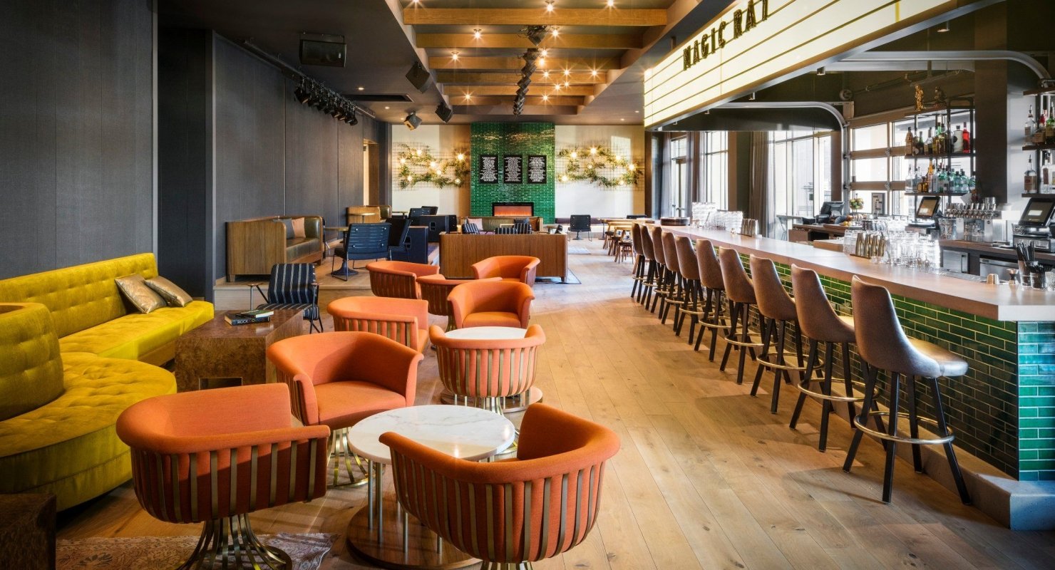 The bar at Magic Rat, a live music venue in the Elizabeth Hotel, featuring warm recessed lighting, a long bar and retro orange chairs around round tables