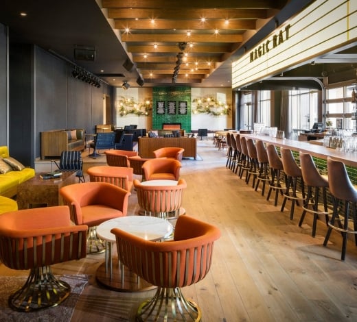 The bar at Magic Rat, a live music venue in the Elizabeth Hotel, featuring warm recessed lighting, a long bar and retro orange chairs around round tables