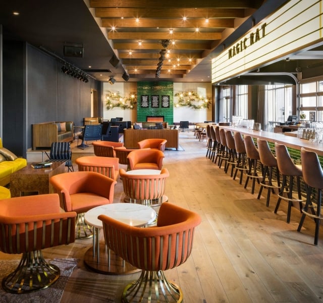 The bar at Magic Rat, a live music venue in the Elizabeth Hotel, featuring warm recessed lighting, a long bar and retro orange chairs around round tables