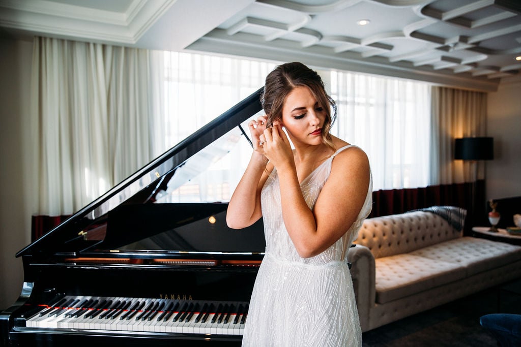 A bride adjust her earring in front of a grand piano as she prepares for her wedding at the Elizabeth Hotel