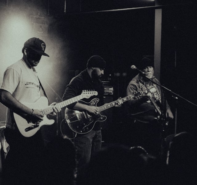 Three guitarists perform on a dimly lit stage, focused on their instruments during a live gig in black and white.