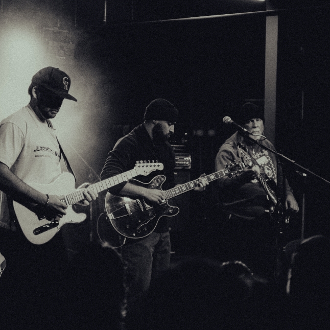 Three guitarists perform on a dimly lit stage, focused on their instruments during a live gig in black and white.