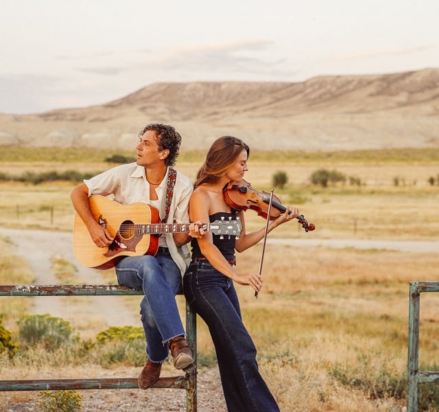 Two musicians, a man with an acoustic guitar and a woman playing a violin, sit on a metal railing in a sunlit desert landscape at golden hour.