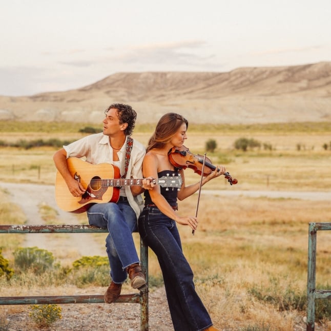 Two musicians, a man with an acoustic guitar and a woman playing a violin, sit on a metal railing in a sunlit desert landscape at golden hour.