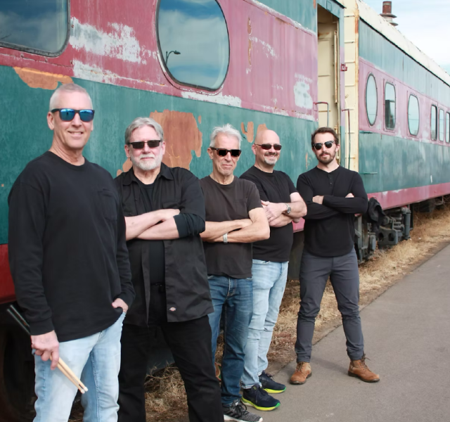 Five men in black shirts and sunglasses stand in a line with arms crossed beside a weathered, turquoise-pink train car on a sunny day.