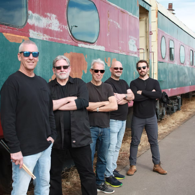 Five men in black shirts and sunglasses stand in a line with arms crossed beside a weathered, turquoise-pink train car on a sunny day.