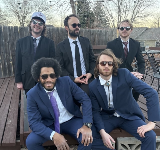 Five men in suits posing outdoors on a wooden deck, some wearing sunglasses; casual group photo.