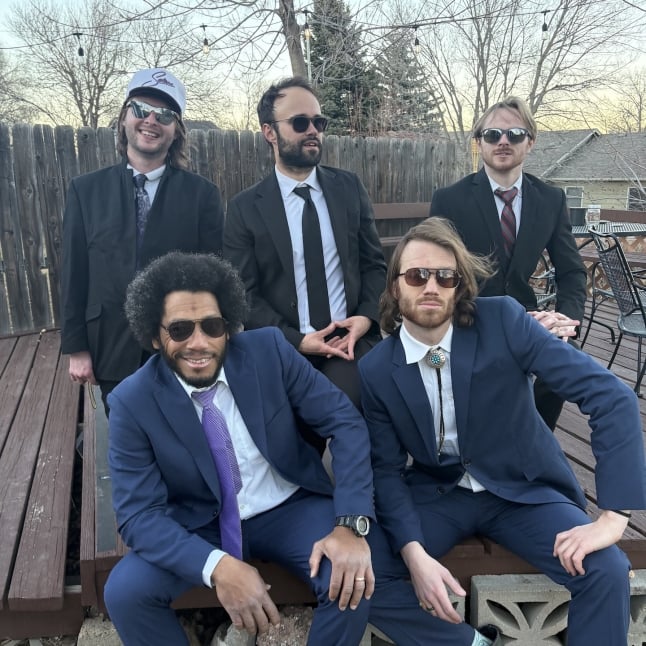 Five men in suits posing outdoors on a wooden deck, some wearing sunglasses; casual group photo.