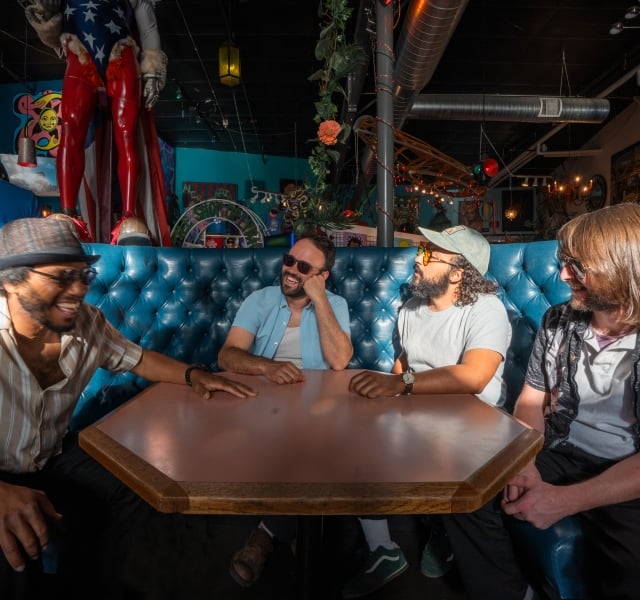 Group of five friends seated on a blue tufted booth around a table, smiling and chatting in a colorful bar interior.