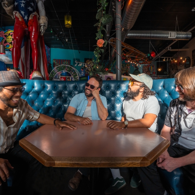Group of five friends seated on a blue tufted booth around a table, smiling and chatting in a colorful bar interior.