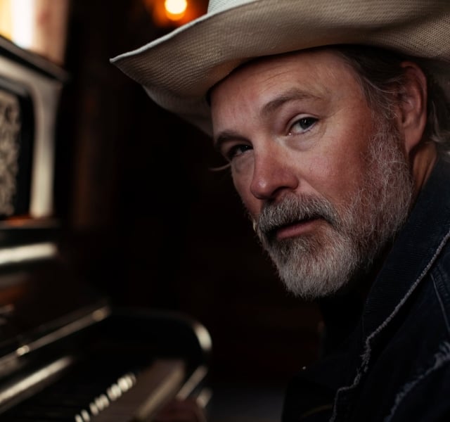 Man with a white hat and gray beard leans beside a piano, looking toward the camera in a warm, dimly lit setting.