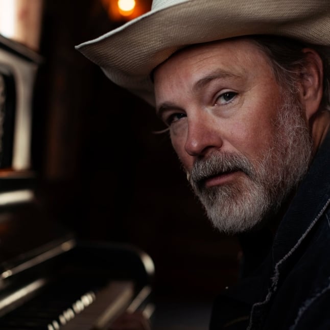 Man with a white hat and gray beard leans beside a piano, looking toward the camera in a warm, dimly lit setting.