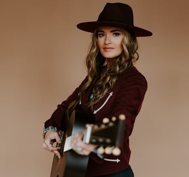 Woman wearing a wide-brimmed hat and maroon jacket playing an acoustic guitar against a beige background.