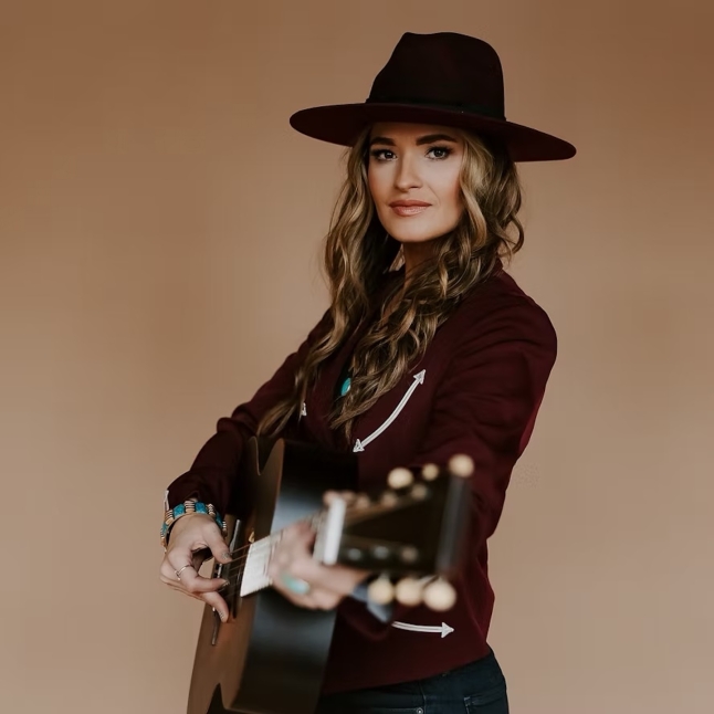 Woman wearing a wide-brimmed hat and maroon jacket playing an acoustic guitar against a beige background.
