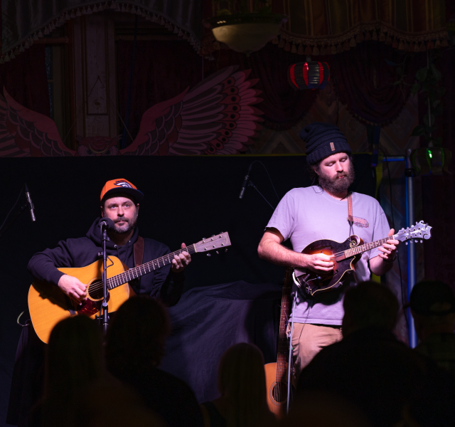 Two musicians perform on a dimly lit stage: one with an orange cap playing an acoustic guitar, the other with a black hat playing a mandolin while singing.