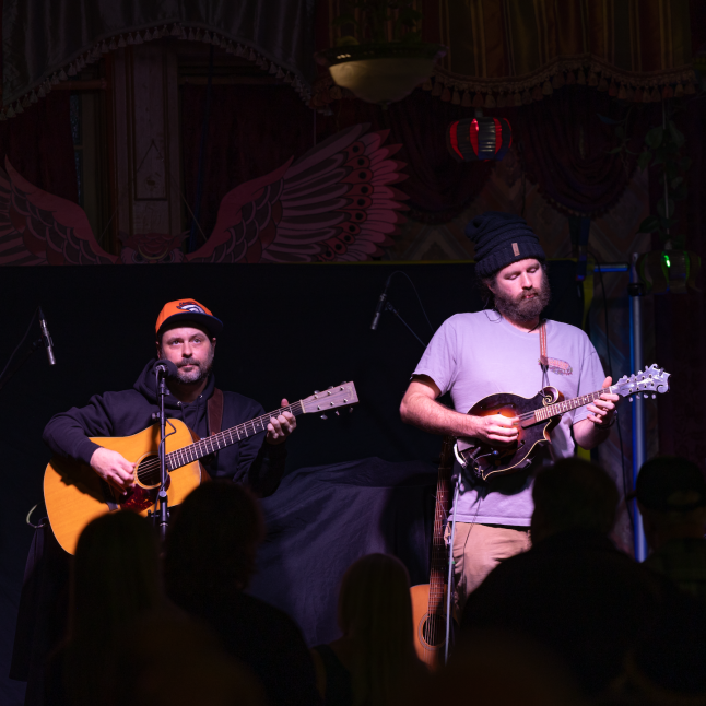 Two musicians perform on a dimly lit stage: one with an orange cap playing an acoustic guitar, the other with a black hat playing a mandolin while singing.