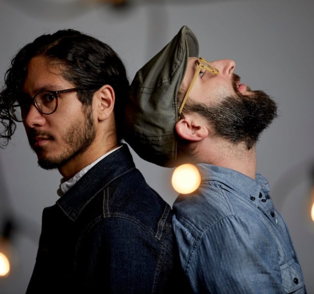 Two men back-to-back in a studio, one with glasses and dark hair, the other wearing a cap and beard, surrounded by warm hanging bulbs.