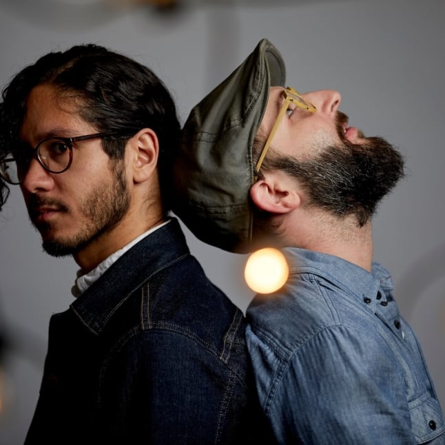 Two men back-to-back in a studio, one with glasses and dark hair, the other wearing a cap and beard, surrounded by warm hanging bulbs.