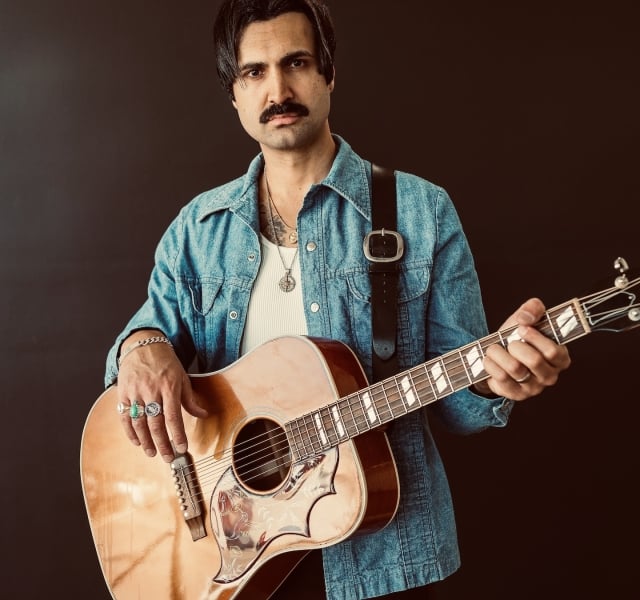 Man with dark hair and a mustache wearing a denim shirt, playing an acoustic guitar against a dark background.