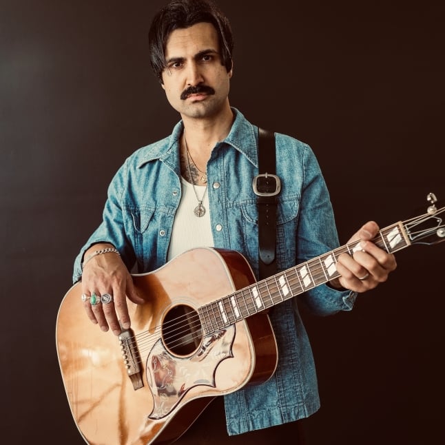 Man with dark hair and a mustache wearing a denim shirt, playing an acoustic guitar against a dark background.