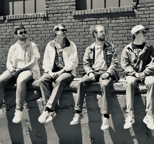 Four men sit on a concrete ledge against a brick wall, wearing casual clothes and sunglasses, looking in different directions.