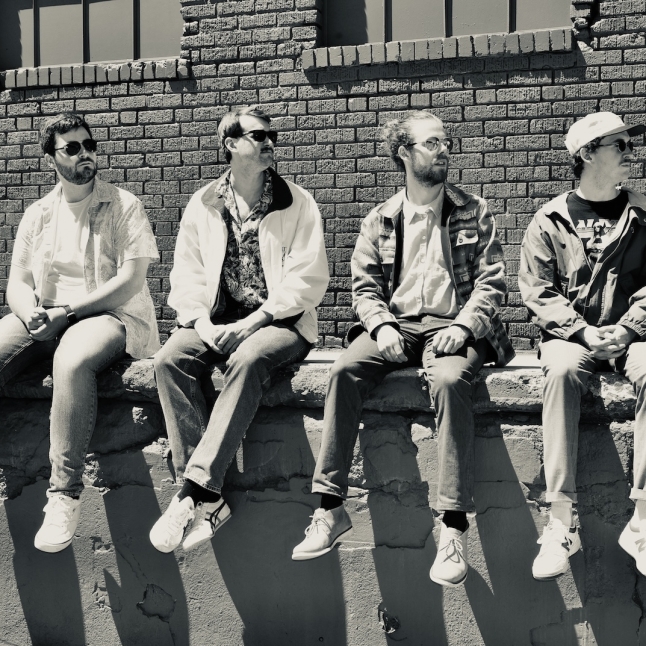 Four men sit on a concrete ledge against a brick wall, wearing casual clothes and sunglasses, looking in different directions.