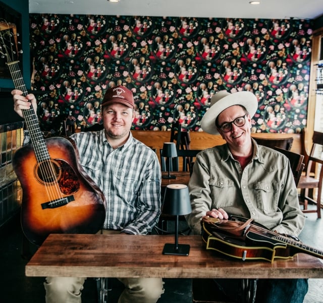 Two men sit at a wooden table in a cafe, one holding an acoustic guitar and the other smiling with a second guitar on the table, colorful floral wallpaper behind them.