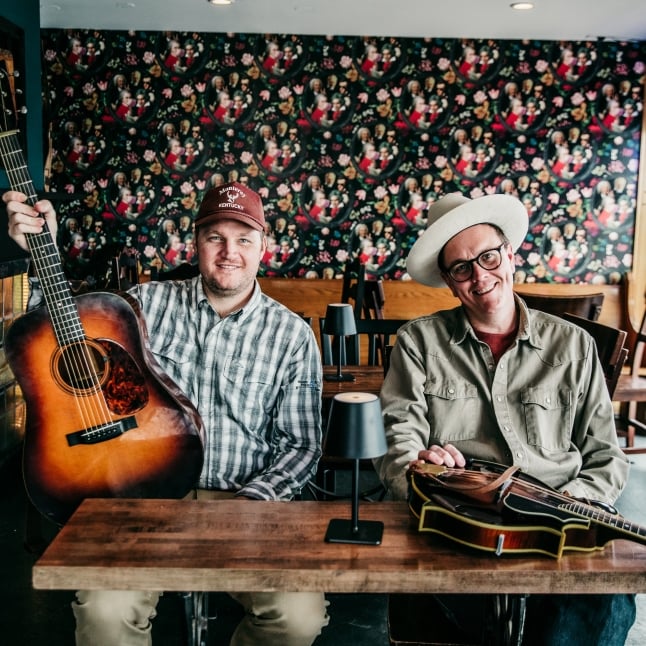 Two men sit at a wooden table in a cafe, one holding an acoustic guitar and the other smiling with a second guitar on the table, colorful floral wallpaper behind them.