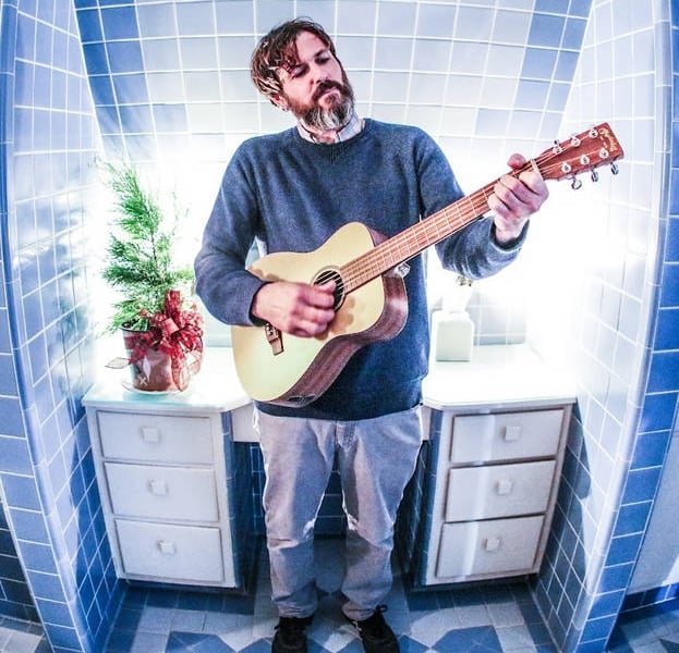 Bearded man in a blue sweater playing an acoustic guitar in a small, blue-tiled bathroom between two white sinks cabinets