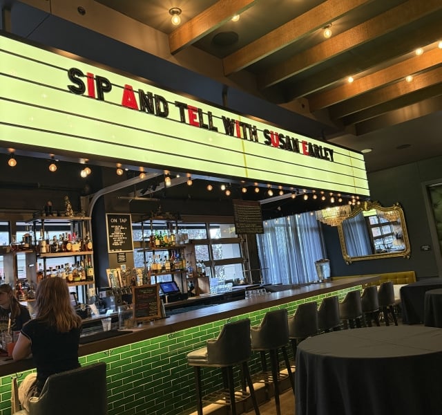 Bar counter with green tile front, row of high stools, and a neon sign reading 'Sip and Tell with Susan' above the bar, illuminated by string lights.