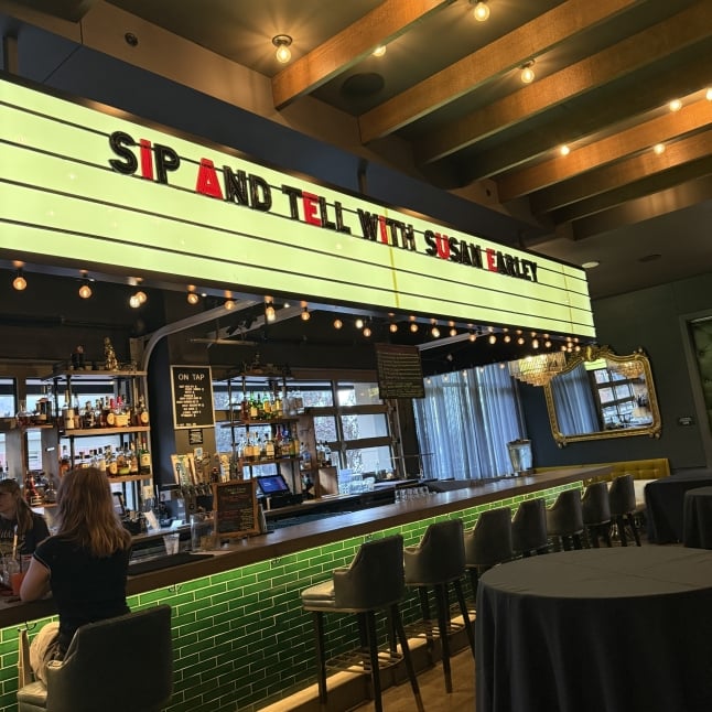 Bar counter with green tile front, row of high stools, and a neon sign reading 'Sip and Tell with Susan' above the bar, illuminated by string lights.