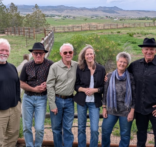 Six adults posing together outdoors by a wooden fence, with green fields and distant mountains in the background.