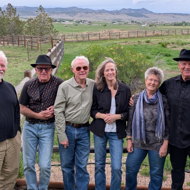 Six adults posing together outdoors by a wooden fence, with green fields and distant mountains in the background.