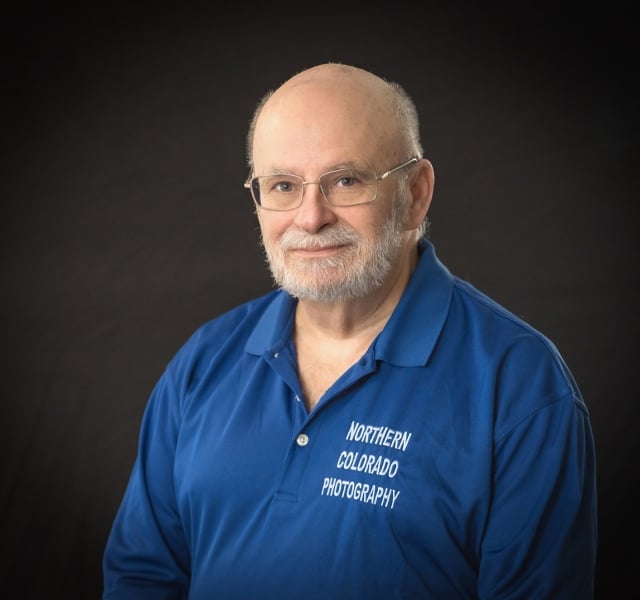 Portrait of an older man with glasses and a white beard, wearing a blue polo shirt that reads 'Northern Colorado Photography' against a dark background.