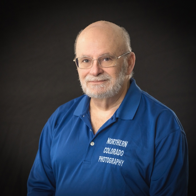 Portrait of an older man with glasses and a white beard, wearing a blue polo shirt that reads 'Northern Colorado Photography' against a dark background.