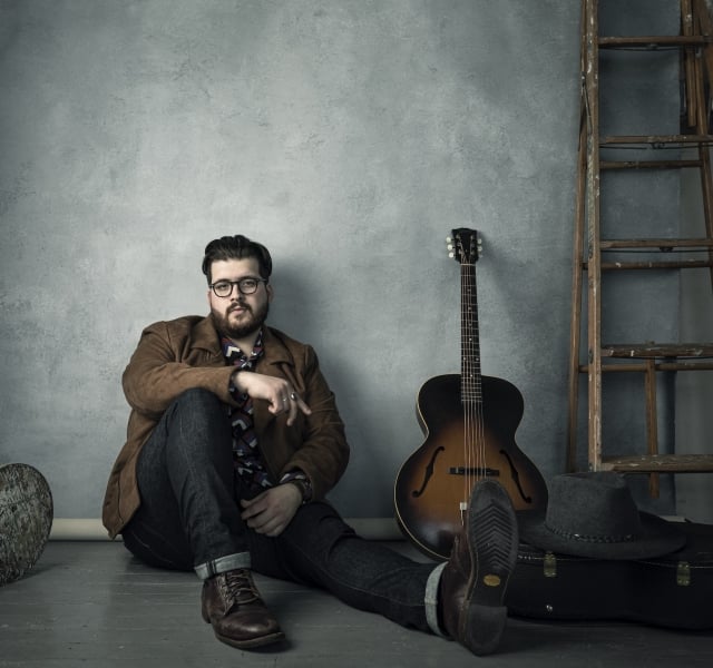 Man with glasses sits on the floor beside a sunburst acoustic guitar in an industrial room with a wooden ladder nearby.
