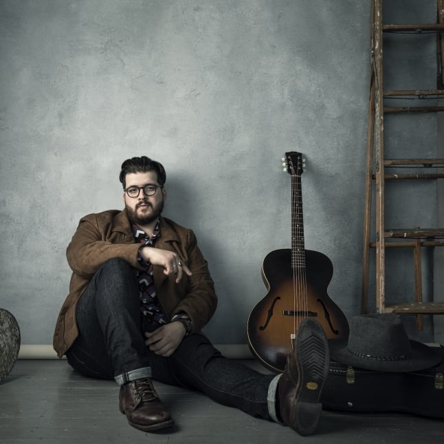 Man with glasses sits on the floor beside a sunburst acoustic guitar in an industrial room with a wooden ladder nearby.